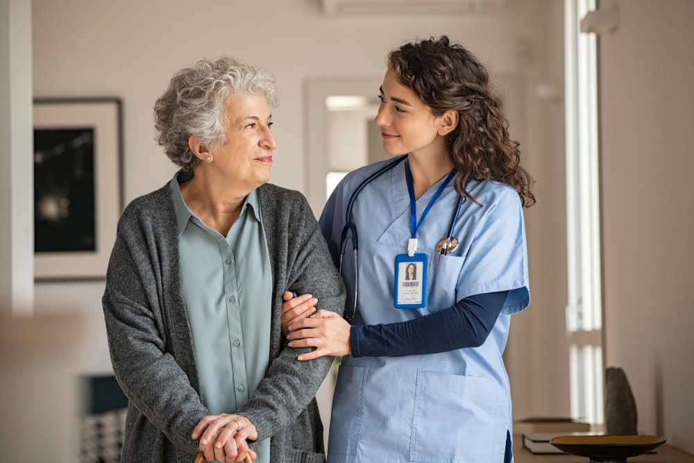 A nurse wearing blue scrubs holding onto a patients arm while they smile and walk through a house