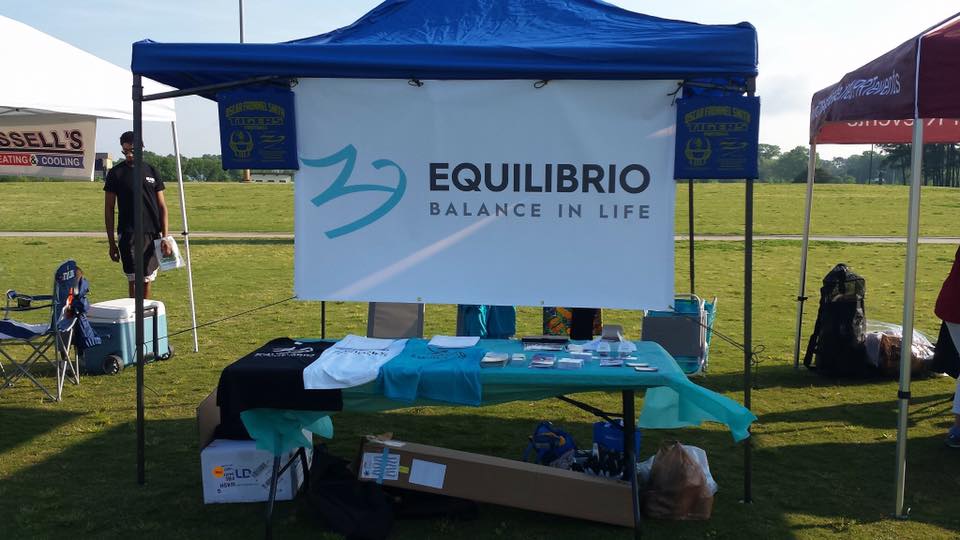 An outdoor booth with a blue canopy displaying a banner that reads "EQUILIBRIO BALANCE IN LIFE." The table beneath holds various items, including a T-shirt, flyers, and small boxes. The booth is set up on green grass with other tents and people in the background.