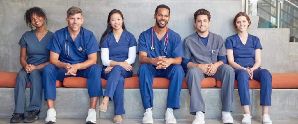 Six medical professionals in scrubs sit on a bench, smiling and facing the camera. The group is diverse, with individuals of different genders and ethnic backgrounds, each wearing a stethoscope or medical uniform. A modern, well-lit hospital corridor forms the background.