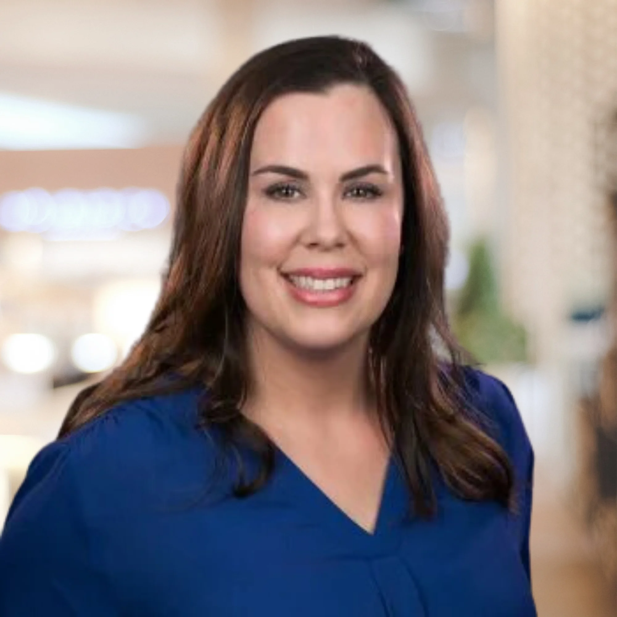 Kimberly Silver Headshot - A woman with long brown hair smiles at the camera in a professional setting. She is wearing a royal blue blouse. The blurred background features soft lighting and neutral colors.
